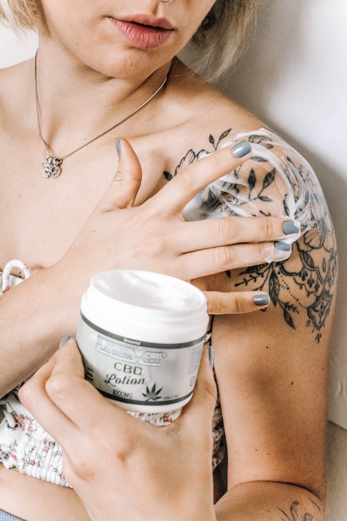 Tattooed woman applying CBD lotion on shoulder for skincare and relaxation.